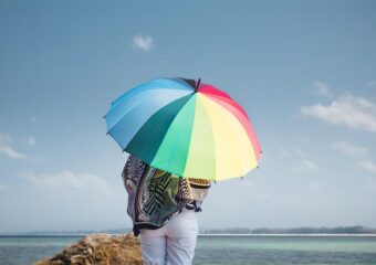 A person holding a rainbow umbrella stands on a rock near the ocean.